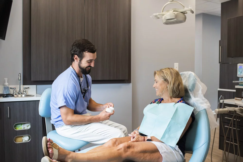 Dr. Jon Byrd of High Point Dental Partners seated on a stool, interacts with a patient in a dental office. The dentist holds a dental model while the patient, seated in a dental chair and wearing a bib, listens attentively, surrounded by dental equipment and cabinetry.