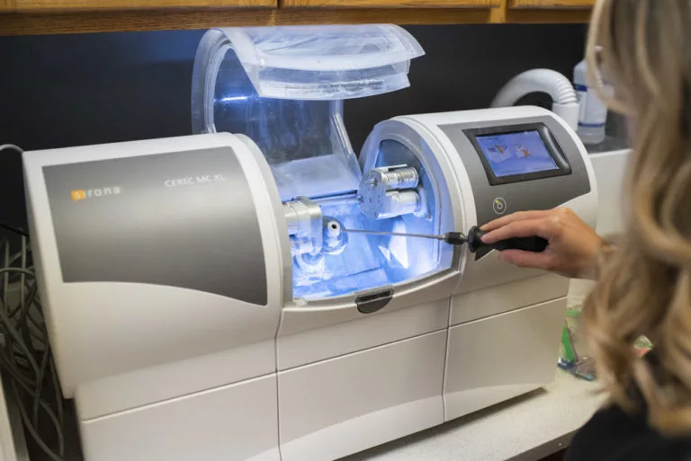 The image shows a CEREC MC XL dental milling machine by Sirona, designed for creating restorations like crowns and veneers. A person interacts with the machine, which features a touchscreen interface and a transparent compartment housing its milling tools.