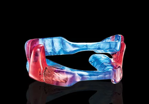 A colorful, translucent dental appliance, primarily blue with red accents, resembling a flexible mouthguard or orthodontic device.