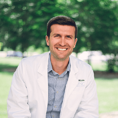 A person in a white lab coat with 'Moore' and 'UNC' embroidered, wearing a light blue shirt, standing outdoors in a green, park-like setting.