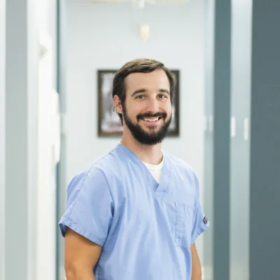 A person wearing light blue medical scrubs standing in a hallway with a framed picture on the wall and several doorways, suggesting a professional medical environment.