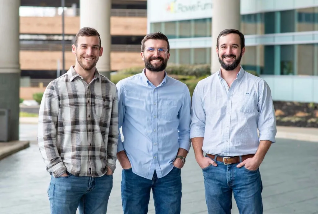 Three individuals standing outdoors; two wearing light blue shirts, one in a plaid shirt. Background shows a modern building with large windows and greenery.