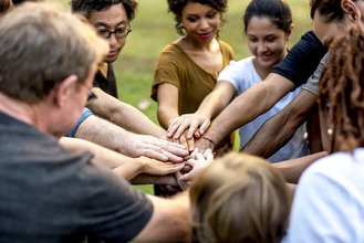 Group of people standing in circle outdoors with hands extended to center, symbolizing teamwork and unity in grassy park setting.
